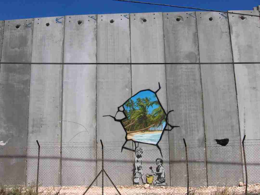 Behind the wall is the beach, Banksy. Image of the West Bank wall with a hole showing the beach, inside the wall some children play with the sand.
