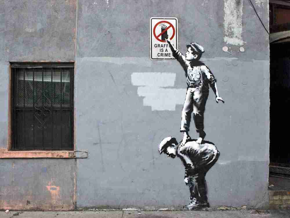 Gaffiti is a crime, Banksy- Mural where two boys appear, one holding the other and the one from arribna tries to collect a spray paint from a sign indicating that graffiti is a crime.